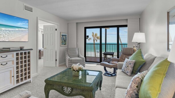 A living room with a glass coffee table, a sofa with cushions, a chair, a television, and a door leading to a balcony with a view of the ocean.