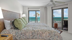A bedroom with a large bed, a ceiling fan, a view of the ocean, and a balcony with a chair.