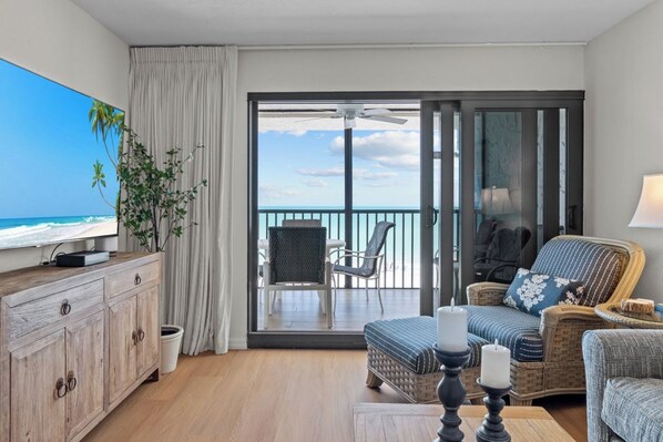 A room with a television, a wooden cabinet, wicker furniture, a balcony with a view of the ocean, and a ceiling fan.