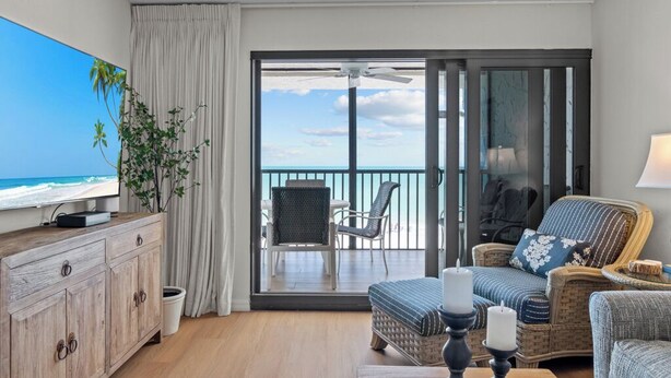 A room with a television, a wooden cabinet, wicker furniture, a balcony with a view of the ocean, and a ceiling fan. - Modern Gulf-Front Condo in Sanibel – Newly Renovated & Ready for Your Escape (Sanibel)
