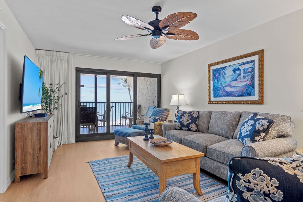 A modern living room with a large flat-screen TV, wooden coffee table, grey sofa with patterned cushions, a ceiling fan, and a large window with a view of the ocean.