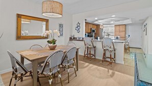 A dining area with a wooden table and patterned chairs, a hanging light fixture, and a kitchen in the background. - Modern Gulf-Front Condo in Sanibel – Newly Renovated & Ready for Your Escape (Sanibel)