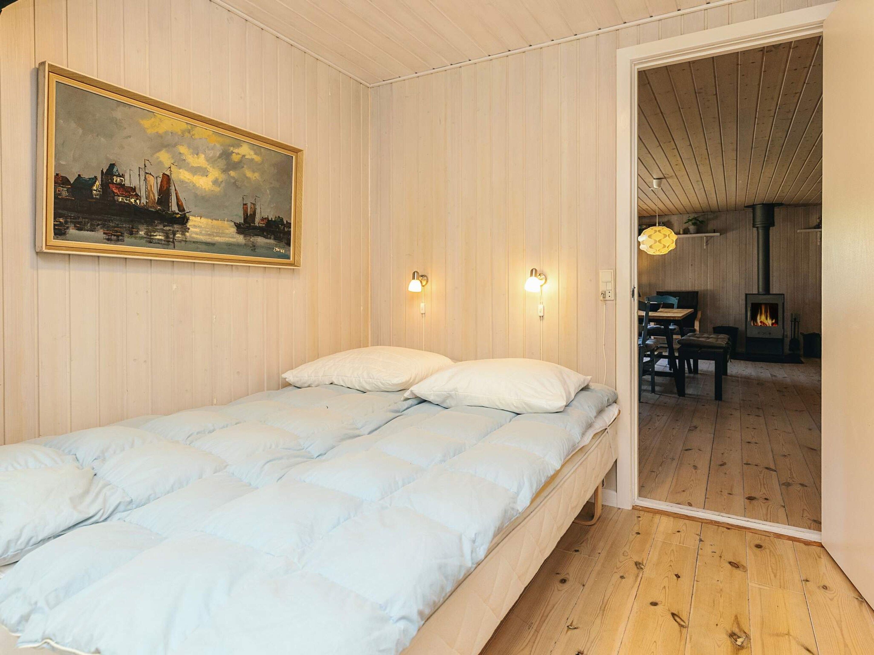 A room with a bed, a painting, two wall-mounted lights, and a visible door leading to another room with a table and chairs.
