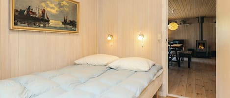 A room with a bed, a painting, two wall-mounted lights, and a visible door leading to another room with a table and chairs.