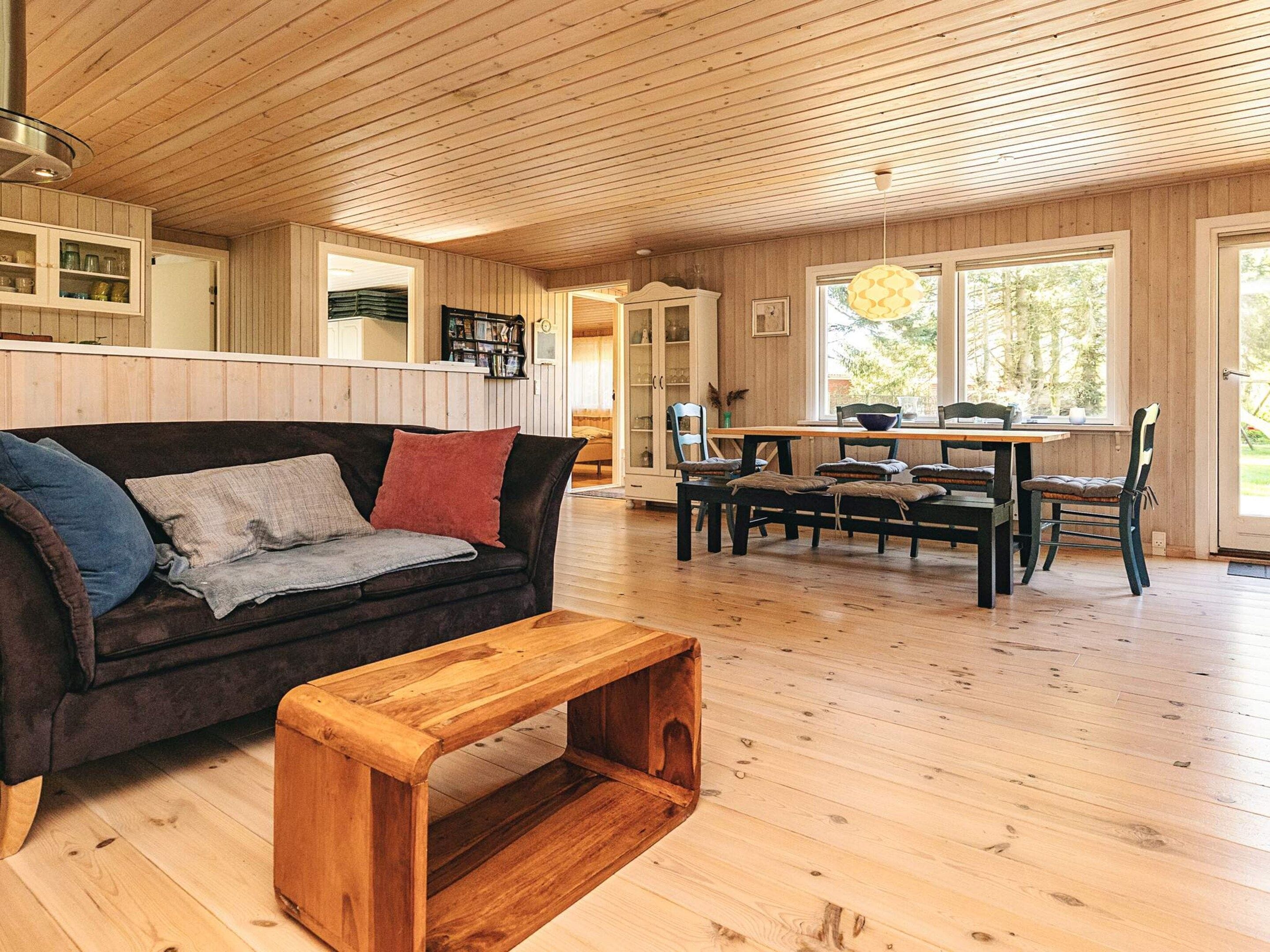 A cozy living room with a wooden ceiling, a dark brown sofa with cushions, a wooden coffee table, and a dining area with a wooden table and chairs.