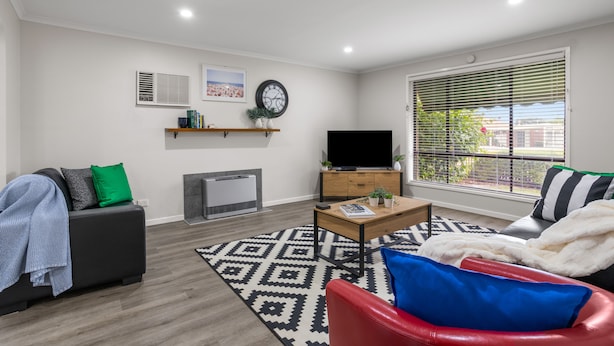 A modern living room with a sofa, a red chair, a coffee table, a flat-screen TV, a fireplace, and a large window with blinds. - Banksia Rosebud  Pet Friendly (Rosebud)