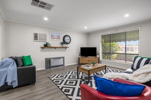 A modern living room with a sofa, a red chair, a coffee table, a flat-screen TV, a fireplace, and a large window with blinds.