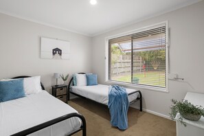 A bedroom with two single beds, a bedside table with a lamp, a framed picture on the wall, and a window with blinds. - Banksia Rosebud  Pet Friendly (Rosebud)