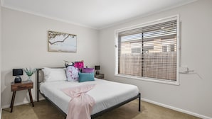 A bedroom with a bed, a nightstand, a lamp, a window with blinds, and a wall-mounted picture. - Banksia Rosebud  Pet Friendly (Rosebud)