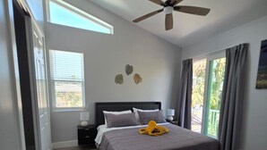 Une chambre à coucher comprenant un lit, un ventilateur de plafond, une table de chevet, une fenêtre avec des stores et une vue sur la verdure à l’extérieur.