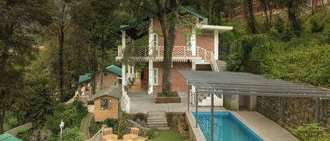 A house with a swimming pool, surrounded by lush greenery and a small garden with outdoor furniture.