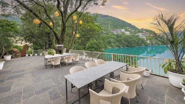 Une terrasse extérieure avec une table et des chaises, entourée de plantes en pot et offrant une vue sur un lac et des collines.
