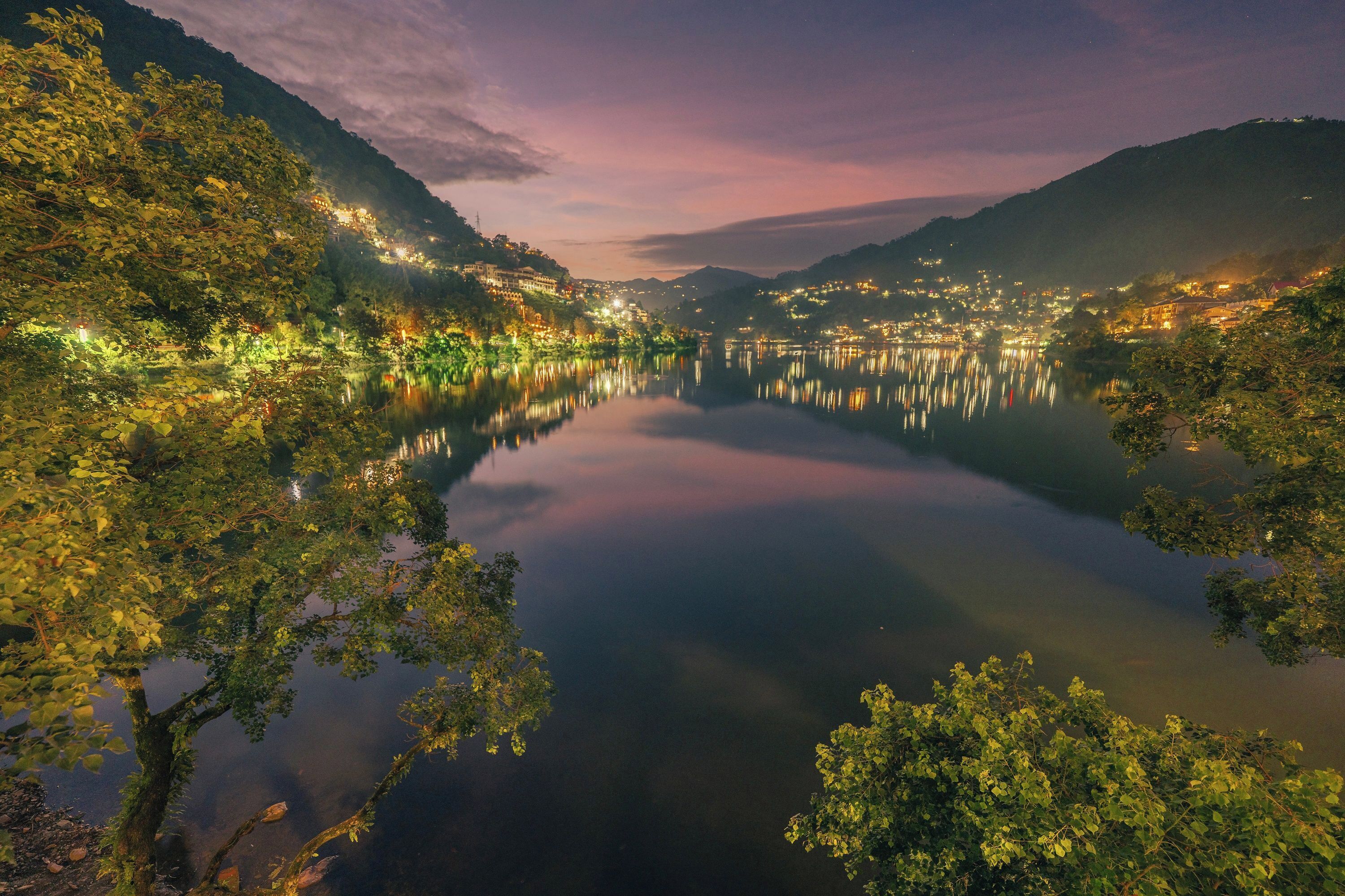 A serene lake surrounded by lush greenery and illuminated buildings, reflecting the lights on the water's surface.