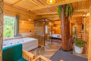 Ein Holzhütten-Innenraum mit Badewanne, einer in Glas eingefassten Dusche, einem Bett und einem großen Baumstamm.