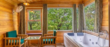 A wooden cabin interior with a wooden floor, a sofa, a table, a bathtub, and large windows with curtains.