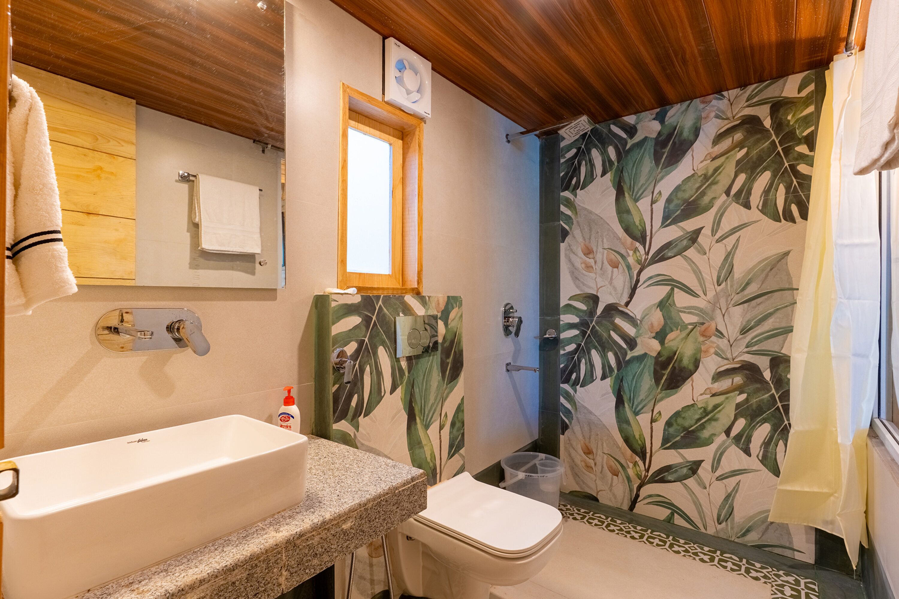A bathroom with a wooden ceiling, a sink, a toilet, a shower with a leaf-patterned curtain, and a window.