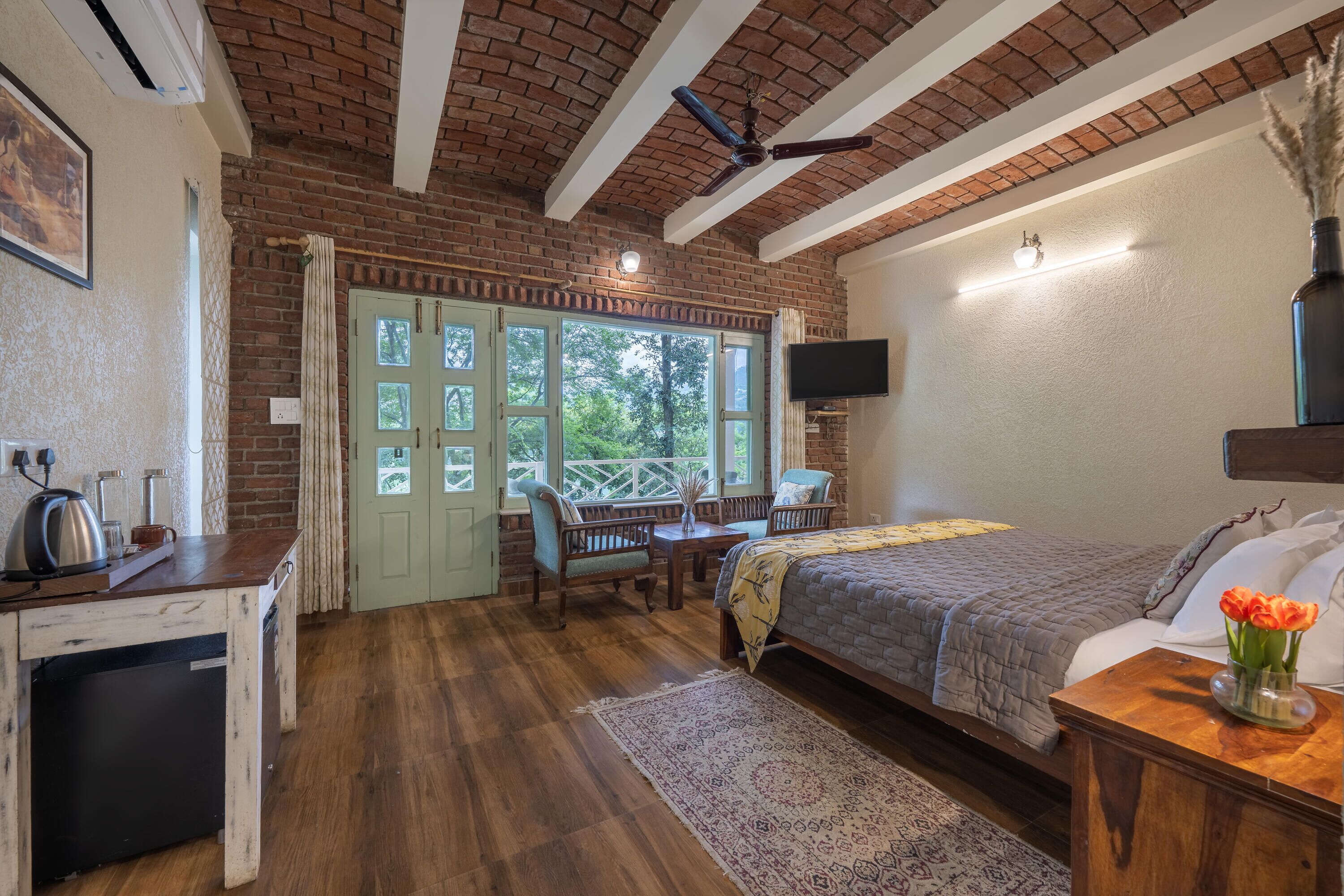 A bedroom with a brick ceiling, a bed with a gray blanket, a wooden nightstand, a TV mounted on the wall, and a window with a view of trees.