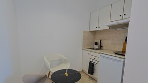 A compact kitchen with white cabinets, a sink, a dishwasher, and a small dining table with a chair. - AVENA Suites (Kato Gouves)