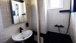 A bathroom with a white sink, a mirror, a shower, and a window. - AVENA Suites (Kato Gouves)