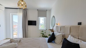 A hotel room with a bed, a television, a window with curtains, a potted plant, and a view of the outside. - AVENA Suites (Kato Gouves)