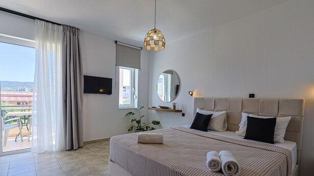 A hotel room with a large bed, a TV, a mirror, and a balcony with a view. - AVENA Suites (Kato Gouves)