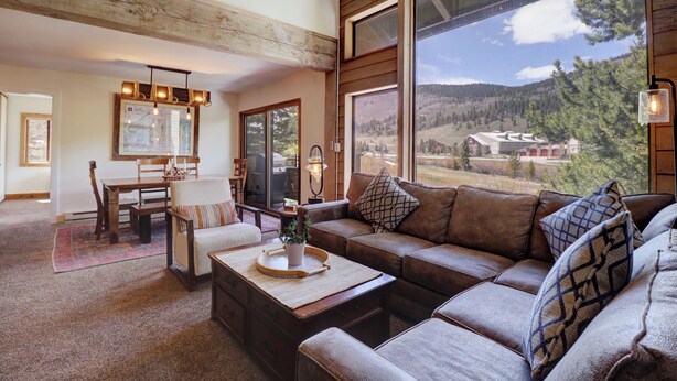 A cozy living room with a large window, a wooden ceiling, and a dining area with a table and chairs.