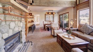 A cozy living room with a stone fireplace, wooden beams, a dining area, and a large window offering a view.