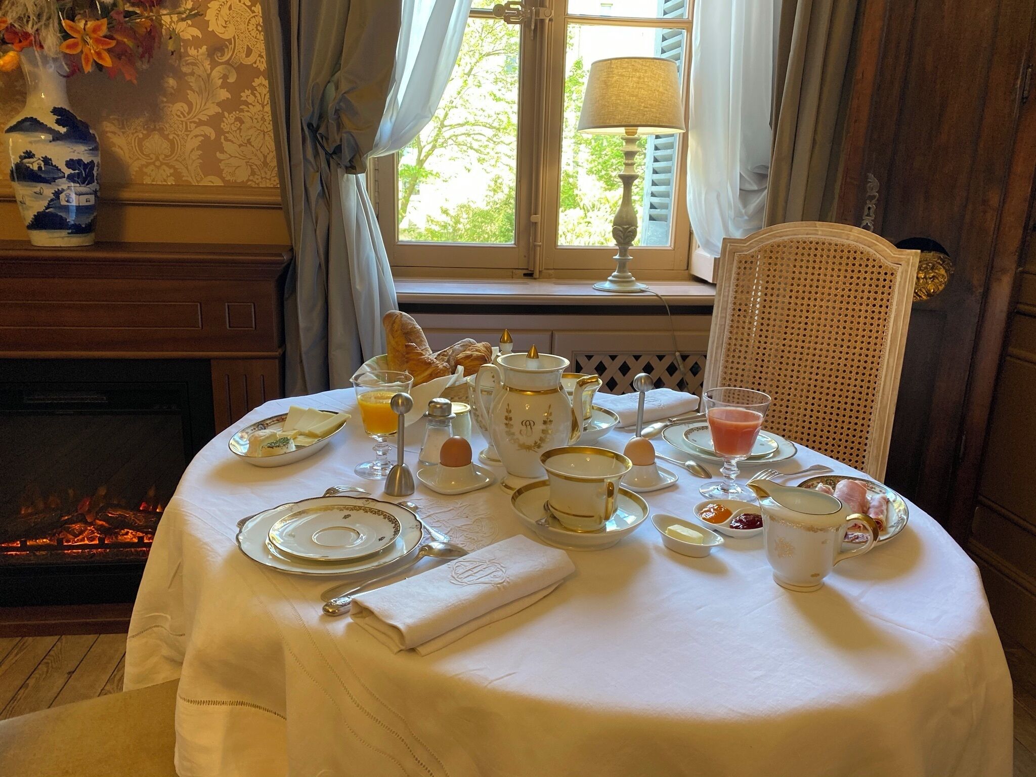 Ein Frühstückstisch ist gedeckt mit Tellern, Tassen und verschiedenen Frühstücksangeboten, darunter Croissants und Orangensaft, in einem Zimmer mit Kamin und einem fenster mit Vorhängen.