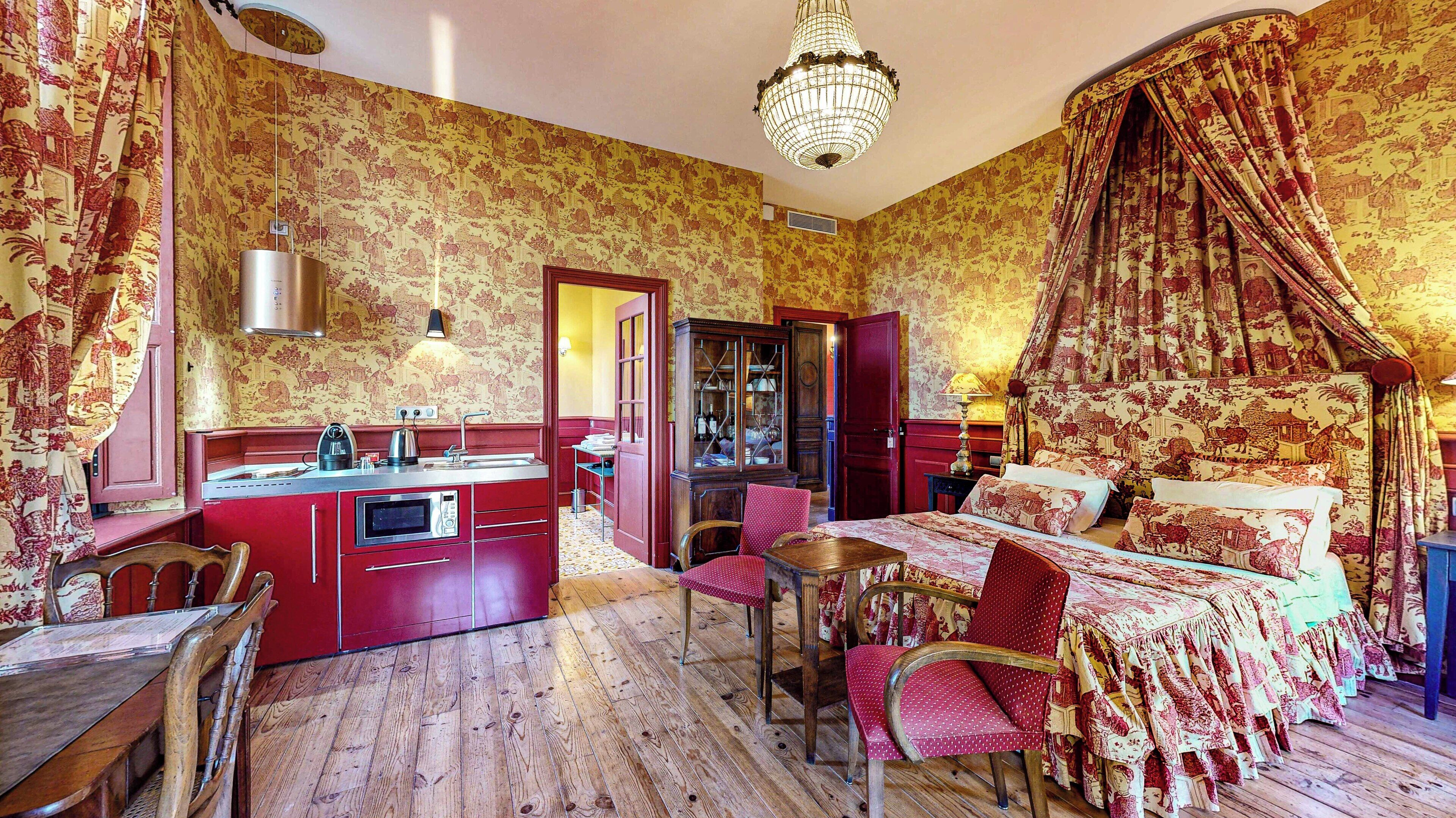 Une pièce dotée d’un plancher en bois, d’un lit à baldaquin, d’une table à manger, de chaises, d’un coin cuisine avec un évier et une cuisinière, et d’un lustre.