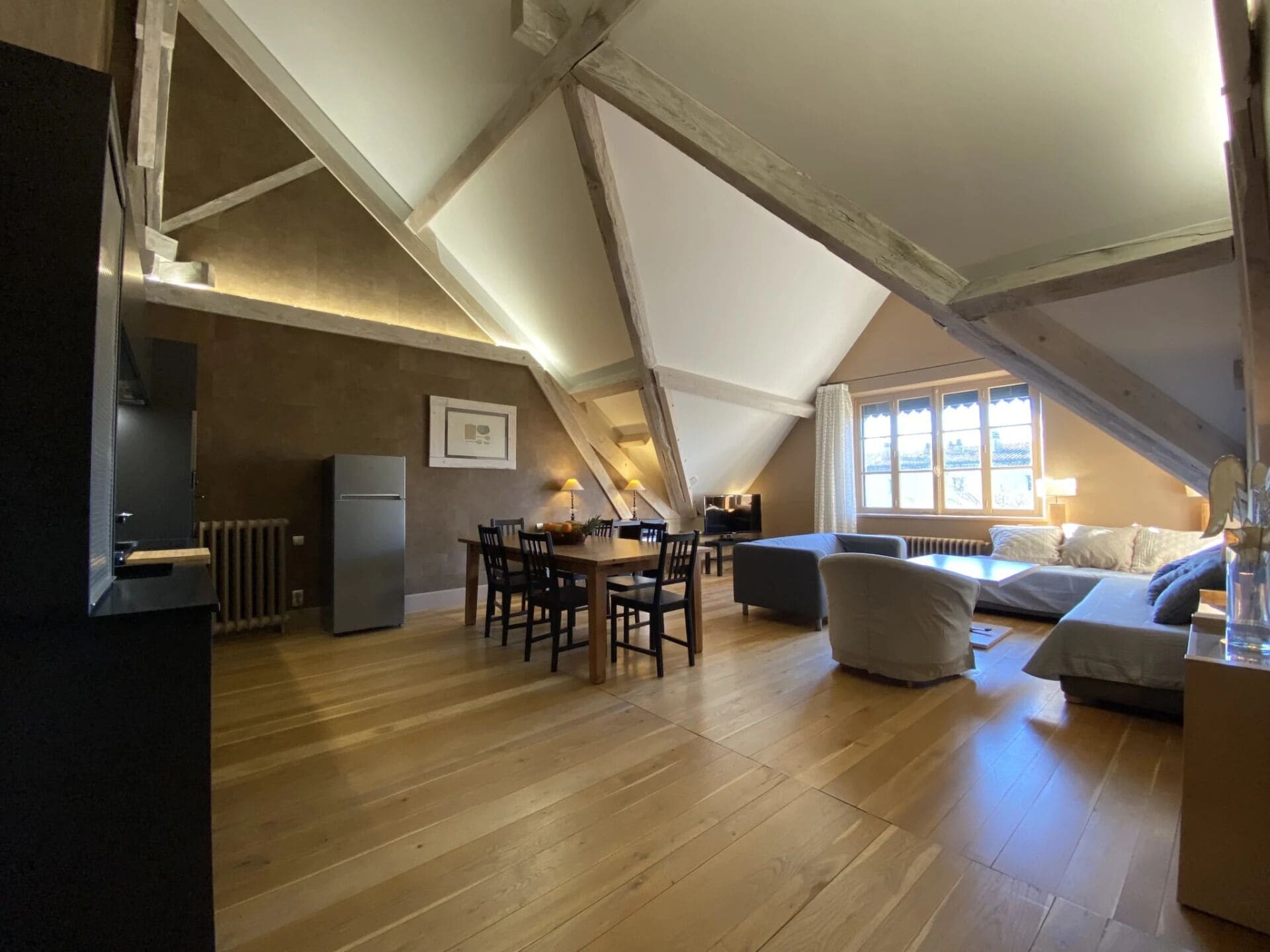 A spacious loft with a high ceiling, exposed wooden beams, a dining area with a table and chairs, a sofa, and a bed.
