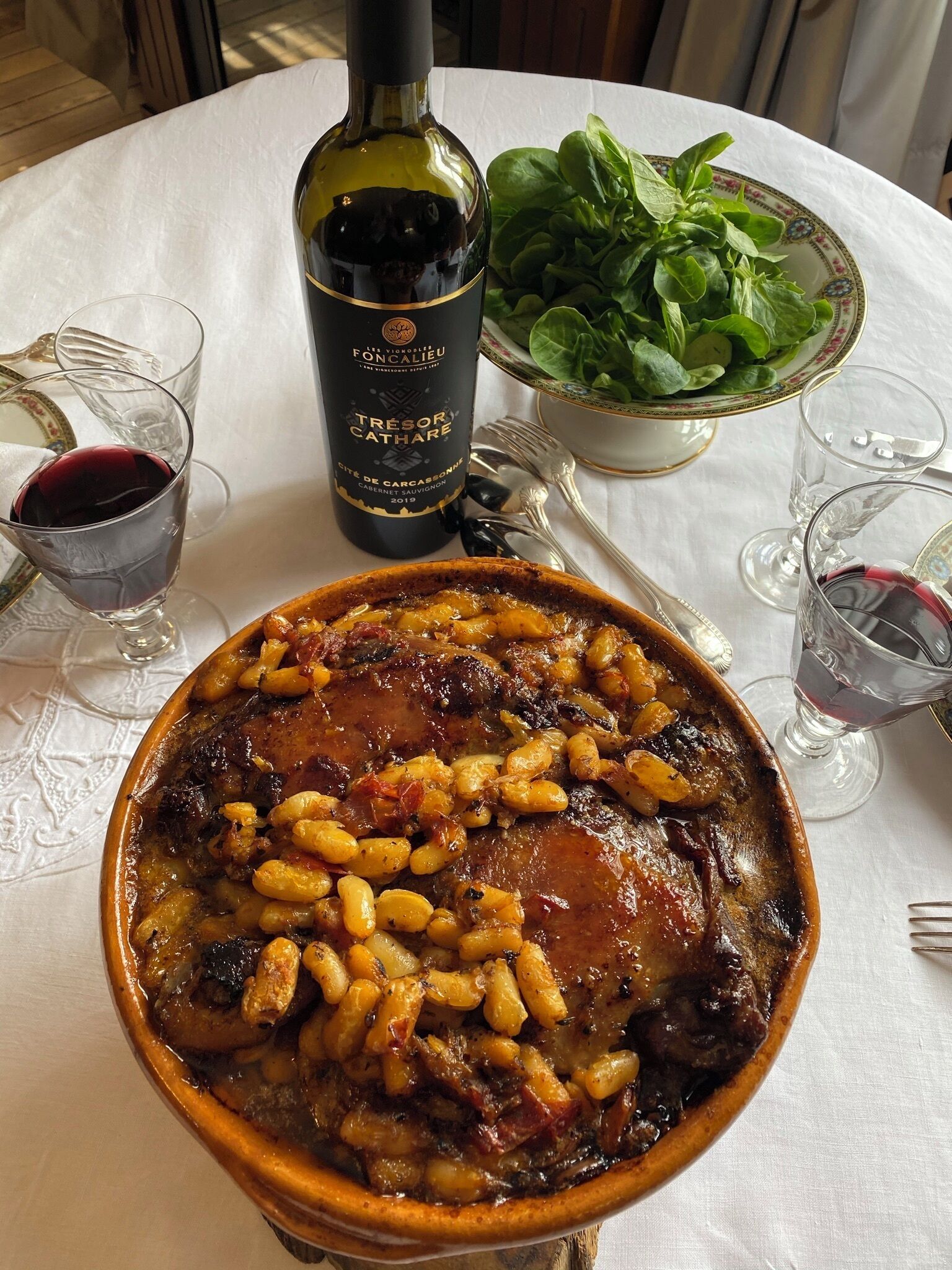 Un repas composé d’une bouteille de vin, d’un saladier de légumes verts et de verres posés sur une table.