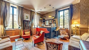 A spacious room with a bar area, multiple seating options including a red armchair, patterned armchair, and a sofa. There are large windows with curtains, a bookshelf, and a decorative mirror. - Demeure Saint Louis (Carcassonne)