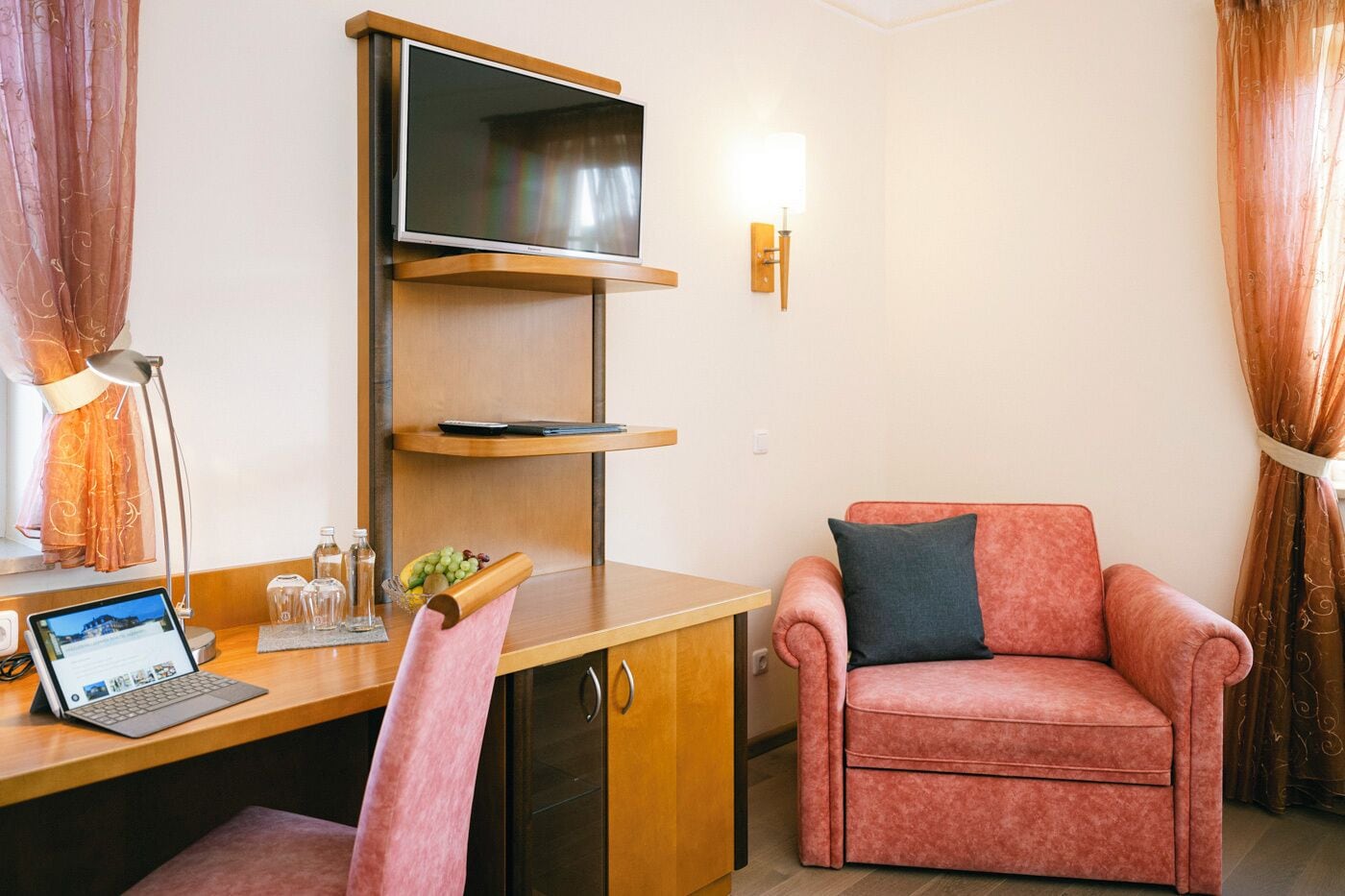 Habitación de hotel compacta con escritorio de madera, una computadora portátil, televisor de pantalla plana, un sillón rosa y una mesita con fruteros.