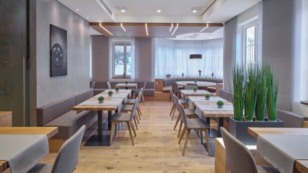 A modern dining area with wooden tables and benches, potted plants, and a large window providing natural light. - Hotel Jägerwirt (Lengau)