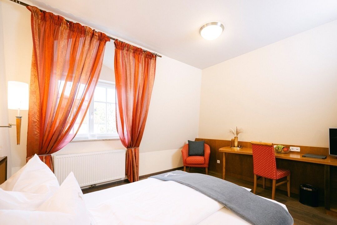 Quarto de hotel com cama, escrivaninha, cadeira e janela com cortinas.
