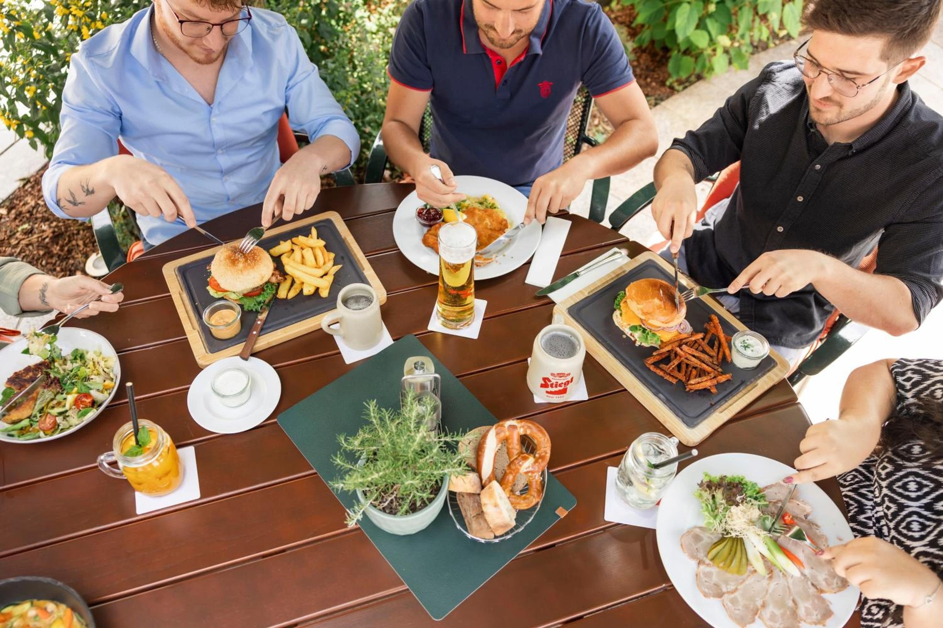 A group of people dining at an outdoor table with various dishes, drinks, and a potted plant.