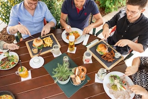 A group of people dining at an outdoor table with various dishes, drinks, and a potted plant.