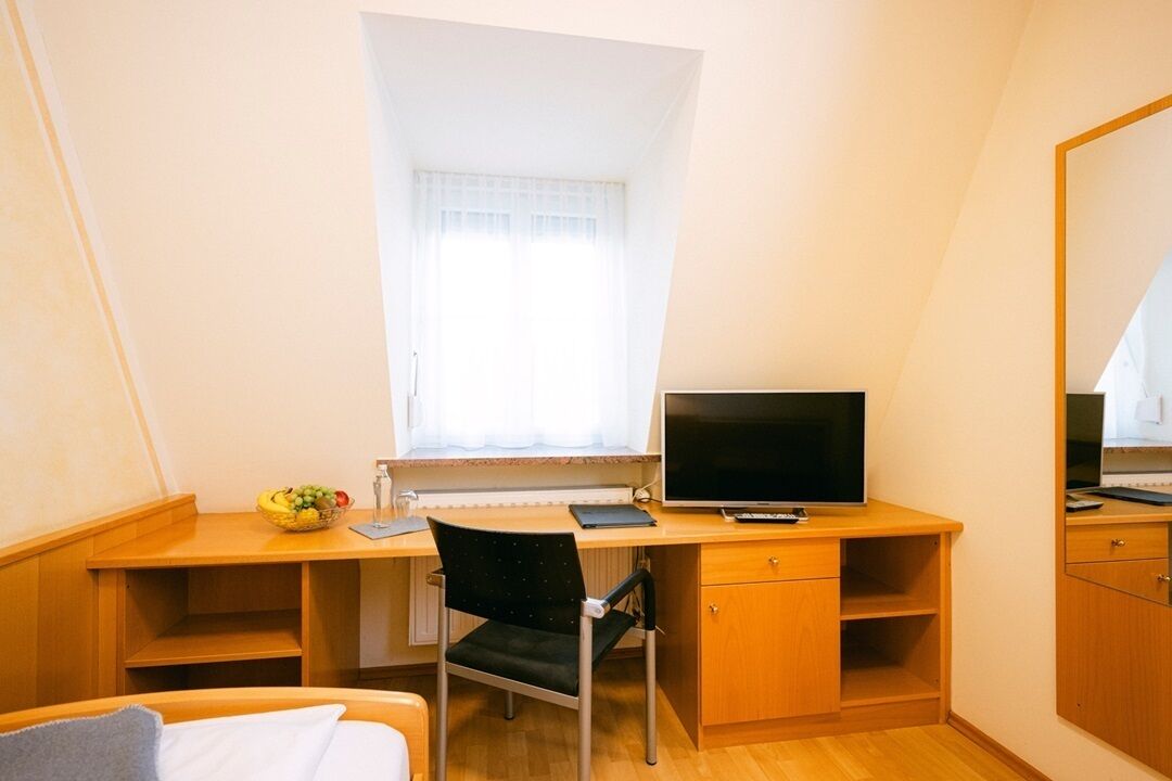 Una habitación pequeña y luminosa con un escritorio de madera, una silla, un televisor y un ventanal con cortinas.