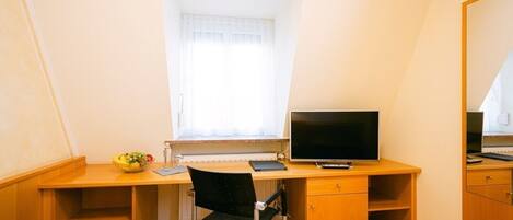 Una habitación pequeña y luminosa con un escritorio de madera, una silla, un televisor y un ventanal con cortinas.
