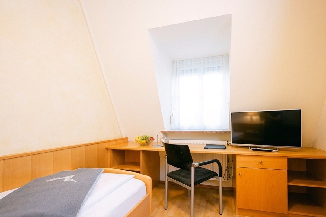 A bedroom with a bed, a desk, a chair, a television, and a window with curtains.