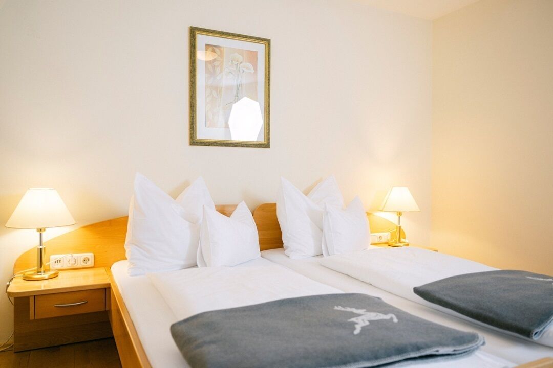 Quarto de hotel com duas camas, roupa de cama branca, cabeceira de madeira, luminárias de cabeceira, mesa de cabeceira e um quadro na parede.
