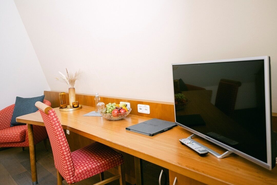 Hotelkamer met een houten bureau, een flatscreen televisie, een laptop, een schaal met fruit en een vaas met gedroogde planten.