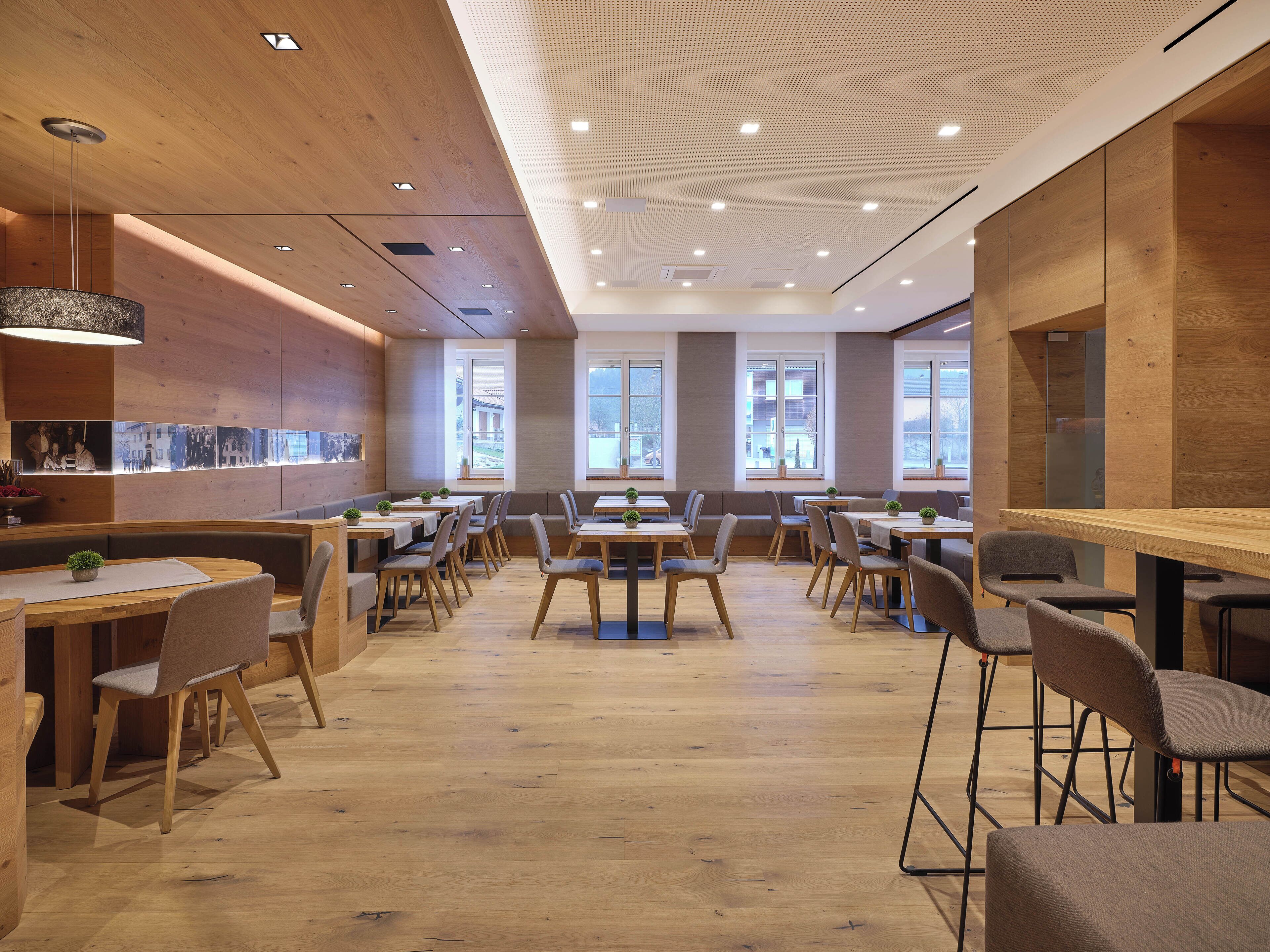 A modern dining area with wooden tables and chairs, large windows, and a ceiling with recessed lighting.