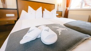 A neatly made bed with white pillows and slippers, a grey blanket with a deer design, and a wooden headboard. - Hotel Jägerwirt (Lengau)
