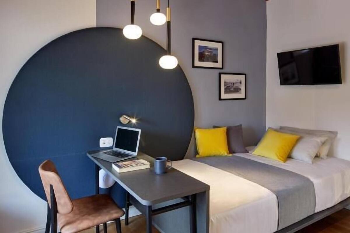 A modern bedroom with a bed, a desk, and a chair. There is a large circular wall feature, a TV, and a ceiling with exposed beams.