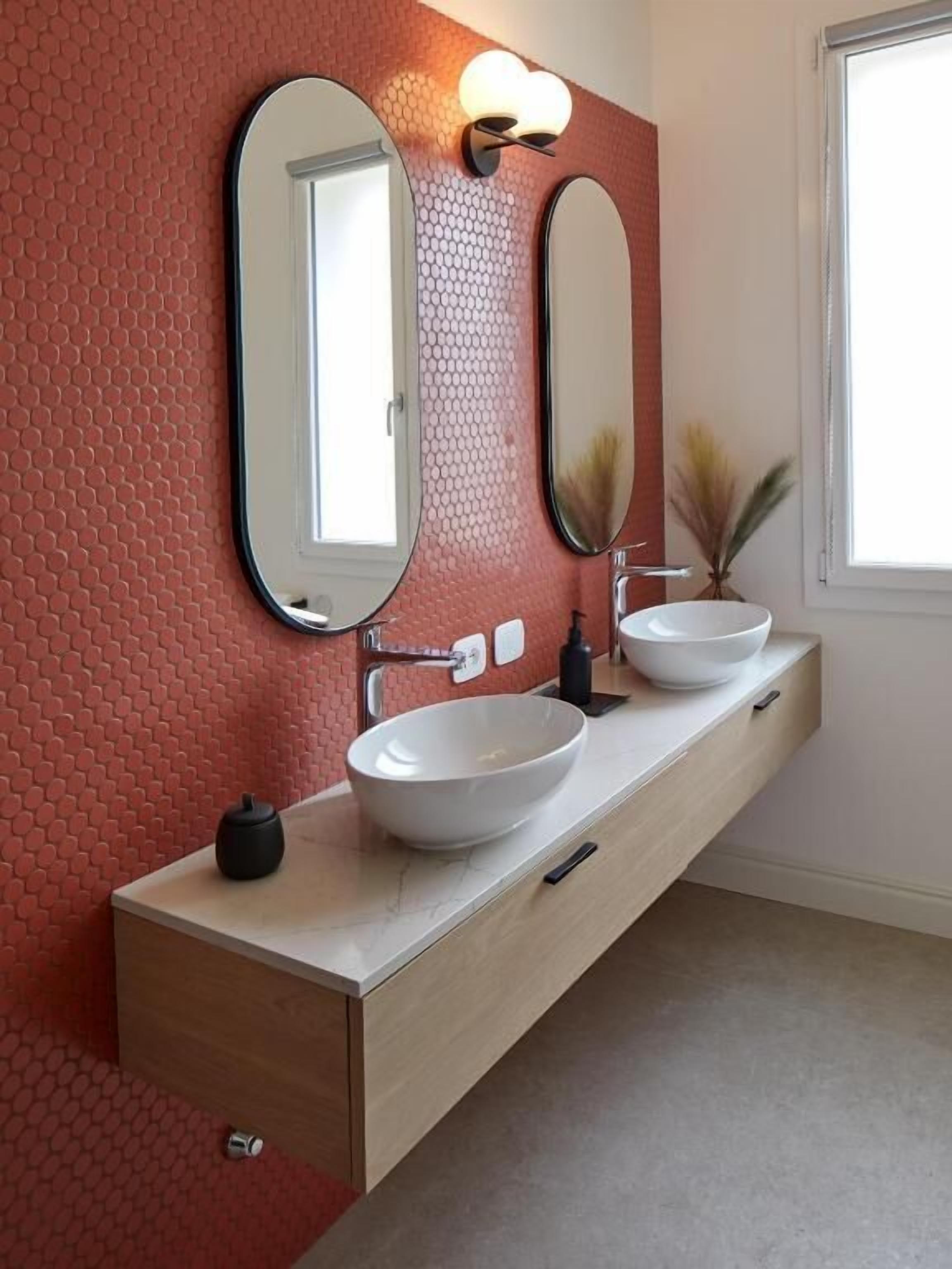 Une salle de bain avec un mur rouge à texture, deux miroirs ovales, un lavabo blanc et un meuble-lavabo en bois.