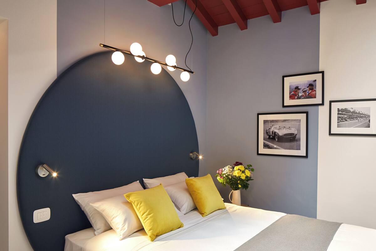 A modern bedroom with a curved headboard, a bed with white and yellow bedding, a bedside table with a vase of flowers, and framed pictures on the wall.