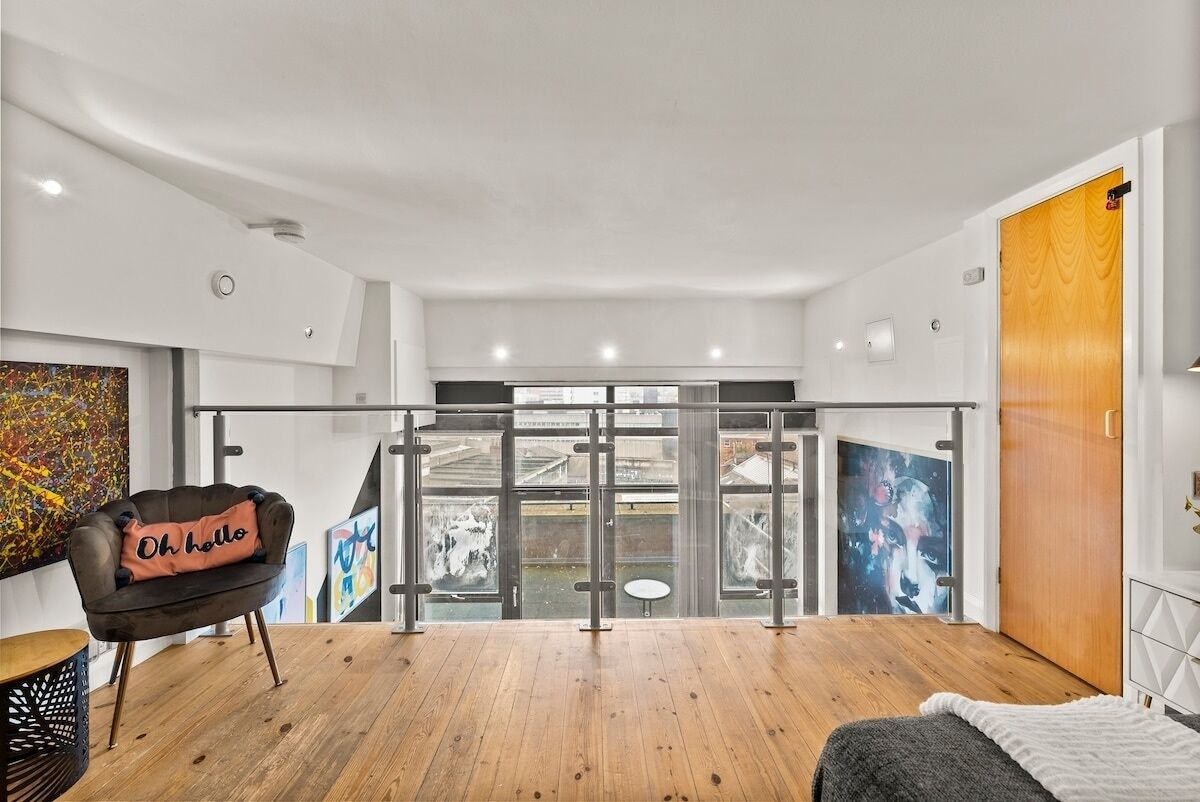 A modern room with a wooden floor, a black chair with a cushion, a bed, a wooden door, and a view through a glass door to the outside.