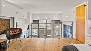 Ein modernes Zimmer mit Holzboden, einem schwarzen Sessel mit Kissen, einem Bett, einer Holztür und Blick durch eine Glastür ins Freie.
