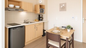 Een compacte keuken met een vaatwasser, magnetron en koffiezetapparaat. Er is een eettafel met stoelen en een kleine potplant.
